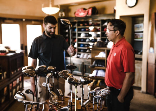 Two men browsing golf clubs in a sports equipment store, discussing options.