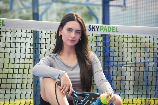 Young woman sitting on padel court, holding racket and ball, looking confident.