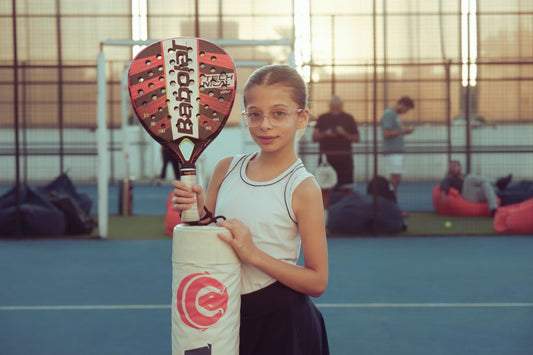 A young girl with glasses holds a paddle racket on an outdoor tennis court during the day.