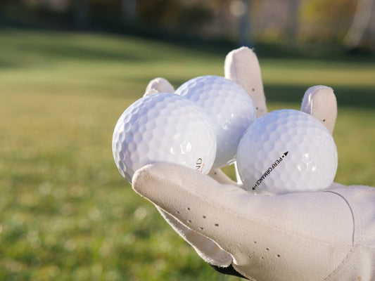 A gloved hand holds three white golf balls on a sunny outdoor golf course.
